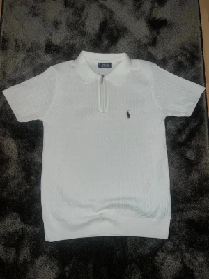 Vit pikétröja med dragkedja Polo Ralph Lauren - Säljer en stilren vit pikétröja från Polo Ralph Lauren med korta ärmar och diskret strukturmönster. Tröjan har klassisk krage, dragkedja framtill och den ikoniska loggan broderad på bröstet. Perfekt till jeans eller shorts.