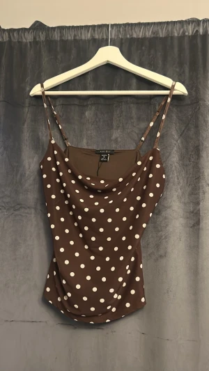 Brun prickig linnetopp - Snygg brun linnetopp med vita prickar och tunna axelband. Toppen har en avslappnad passform med draperad urringning och är perfekt för varma dagar. Materialet känns mjukt och lätt mot huden.