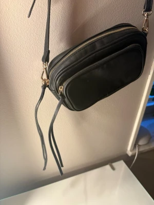 Svart axelväska i skinnimitation - Snygg svart axelväska med två fack, ett stort och ett lite mindre och guldfärgade och fransiga dragkedjor. Väskan har justerbar axelrem och långa dragkedjedetaljer. ❣️ 