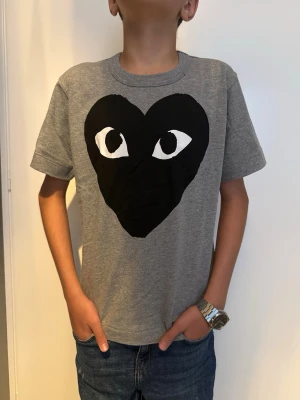 Grå t-shirt med svart hjärta - Snygg grå t-shirt med ett stort svart hjärta och ögon på bröstet. T-shirten har rund halsringning och korta ärmar. 