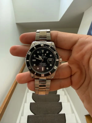 Rolx - Stilren klocka i silverfärgat metallarmband och svart urtavla med tydliga vita markeringar och datumvisning. Klockan har en rund form och vridbar bezel, perfekt för dig som gillar en tidlös och sportig look.