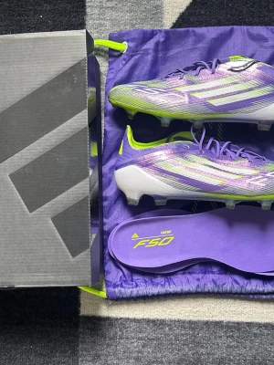 Lila Adidas F50 fotbollsskor - Säljer ett par snygga Adidas F50 fotbollsskor i lila och vitt med neon-gula detaljer. Skorna har en modern, strömlinjeformad design och levereras med matchande skosäck och extra innersula. Perfekta för dig som vill sticka ut på planen.