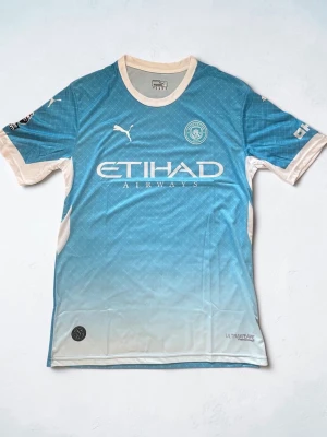 Manchester City matchtröja Puma - Snygg ljusblå Manchester City matchtröja från Puma med vita detaljer och klubbmärke på bröstet. Tröjan har rund halsringning, korta ärmar och tryckt sponsorlogga framtill. Perfekt för fotbollsträning eller att bära till vardags.