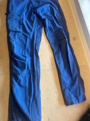 Blå cargobyxor med fickor - Snygga blå cargobyxor med flera praktiska fickor på benen och avslappnad passform. Tillverkade i ett slitstarkt tyg som passar perfekt för en casual och street-inspirerad stil. Perfekta för dig som gillar funktionella och trendiga byxor. Samt att ha när man är ute och vistas i skogen!