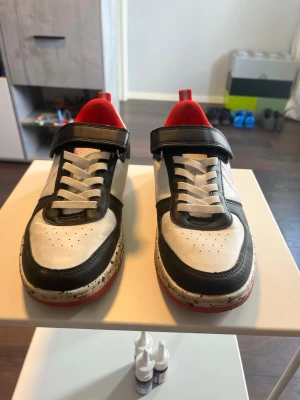 Svarta och vita sneakers med röd sula - Snygga sneakers i svart och vitt med röda detaljer och röd insida. Skorna har vita snören, kardborreband och en cool stänkmönstrad sula. Materialet är en mix av läder och syntet. Perfekta för dig som vill sticka ut med din stil.