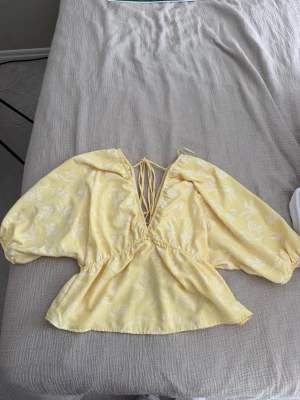 Gul blommig blus med puffärmar - Söt gul blus med vita blommönster och djup v-ringning. Blusen har puffiga trekvartsärmar och knytband bak. 