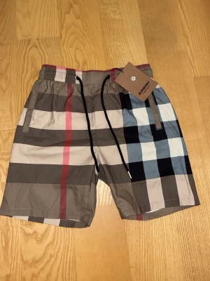 Rutig Burberry shorts med snörning - Står M på tagsen men passar som S.            Snygga rutiga shorts från Burberry med klassiskt mönster i beige, svart, vit och röd. Shortsen har elastisk midja med svart snörning och sidofickor. Perfekta för sommaren och ger en trendig look.