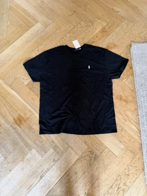 Svart t-shirt med liten logga extra large - En klassisk svart t-shirt med rund hals och korta ärmar. På bröstet finns en liten broderad logga i vitt som ger en stilren touch. Perfekt basplagg till garderoben och enkel att matcha med allt.