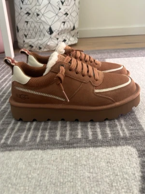 Bruna sneakers från UGG med fuskpäls - Säljer ett par bruna sneakers från UGG med chunky sula och vita detaljer. Skorna har snörning, ovandel i mocka och textil samt mjukt foder i fuskpäls. Perfekta för dig som vill ha både stil och komfort i vardagen.