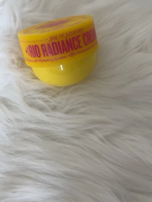 Sol De Janeiro - Sol de Janeiro Rio Radiance Cream är en kroppskräm i en rund, gul burk med lila text. Krämen har Cheirosa 87-doft och innehåller mica för en lystergivande effekt. Perfekt för dig som vill ha glow och återfuktning i ett snyggt förpackat format.