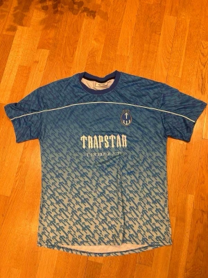 Trapstar t-shirt - Säljer en blå t-shirt från Trapstar med vit text och nummer 22 på ryggen. T-shirten har ett unikt mönster över hela plagget, rund halsringning och korta ärmar. Perfekt för dig som vill sticka ut med streetwear-stil.
