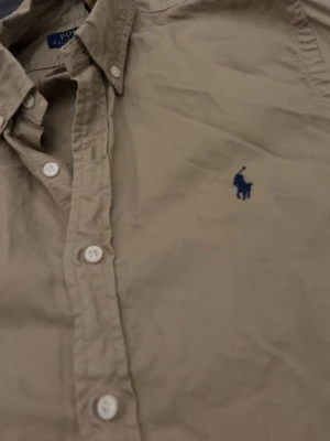 Beige skjorta från Polo Ralph Lauren - Säljer en beige skjorta från Polo Ralph Lauren med klassisk button-down krage och blå broderad logga på bröstet. Skjortan har långa ärmar och vita knappar framtill. Perfekt till jeans eller chinos för en clean look.