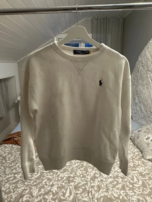 Vit sweatshirt från Polo Ralph Lauren - Vit sweatshirt från Polo Ralph Lauren med rund halsringning och broderad logga på bröstet. Tröjan har ribbade muddar vid ärmslut och nederkant, samt klassisk relaxed passform. Perfekt för en stilren och avslappnad look. Skavanker finns, som hål och missfärgningar (se bilderna) 