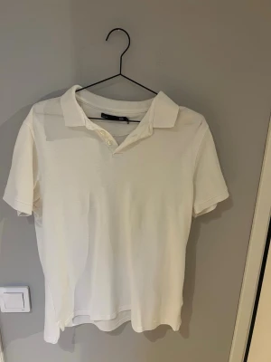Vit pikétröja  - Klassisk vit pikétröja fmed krage och knappar framtill. Tröjan har korta ärmar och är tillverkad i ett mjukt bomullsmaterial. Perfekt för en clean och stilren look.