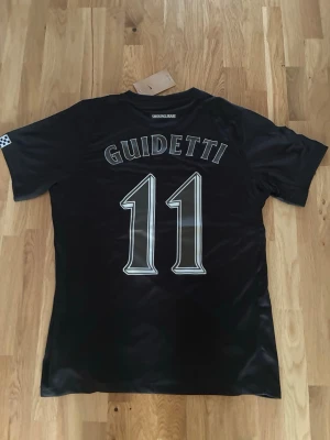 Svart AIK matchtröja Nike Guidetti - Snygg svart AIK matchtröja från Nike med GUIDETTI och nummer 11 på ryggen. Framsidan har AIK:s klubbmärke och årtalet 1891 tryckt i vitt. Tillverkad i ett glansigt, snabbtorkande material som passar perfekt till fotbollsträning eller match.
