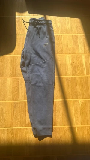 Mörkblå nike tech byxor  - Snygga mörkblå mjukisbyxor från Nike med dragsko i midjan och ribbade muddar vid bensluten. Byxorna har två sidofickor och är tillverkade i mjukt, bekvämt material. Perfekta för chill dagar eller träning.