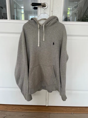 Ralph lauren hoodie - Säljer en grå hoodie med huva och vita dragsnören. Klassisk känguruficka framtill och en liten broderad logga på bröstet. Hoodien har ribbade muddar vid ärmslut och nederkant. Perfekt för en chill och avslappnad stil. Vid fler frågor eller bilder är det bara att skriva