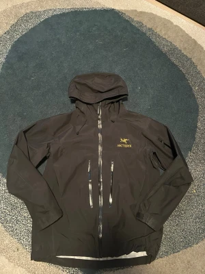 Svart regnjacka från Arc'teryx - Snygg svart regnjacka från Arc'teryx med huva, tejpade sömmar och två stora dragkedjeförsedda fickor framtill. Jackan har justerbar nederkant och logga i gult på bröstet. Perfekt för regniga dagar och outdoor-äventyr.