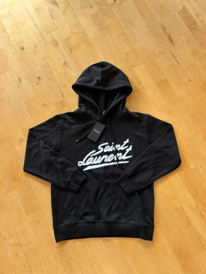 Svart Saint Laurent hoodie med tryck - Snygg svart hoodie från Saint Laurent med vit logga i graffiti-stil på bröstet. Klassisk känguruficka framtill och justerbar huva med snören. Tillverkad i mjukt material som känns skönt mot huden. Perfekt för en avslappnad och trendig look.