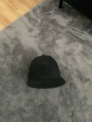 Svart New York Yankees keps - Säljer en svart New York Yankees keps från New Era, modell 59FIFTY. Kepsen har en böjd skärm och broderad NY-logga framtill i samma färg som kepsen. Klassisk streetwear-stil och passar perfekt till en avslappnad look. 55,8cm storlek 7
