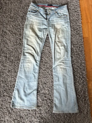 Lågmidjade bootcut jeans Tommy Hilfiger - Säljer ett par ljusblå lågmidjade bootcut jeans från Tommy Hilfiger. Jeansen har klassisk femficksdesign, låg midja och snygga slitningar framtill. Tillverkade i mjukt denimtyg med röda detaljer på insidan av linningen. Har behövt sytt innanför låret pga hål. Storlek 14 och passar på mig som S/xs