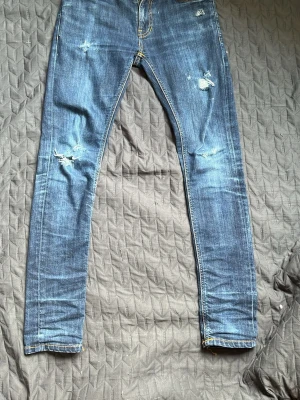 Blå nudie jeans - Snygga blå salvage nudie jeans med slitningar på knäna och låret. Jeansen har  klassiska bakfickor med dekorativa sömmar och är perfekta för en cool och avslappnad archive stil. 