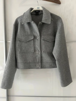 Grå kort jacka med fiskbensmönster - Stilren grå jacka i kort modell med fiskbensmönster och stora knappar framtill. Jackan har klassisk krage, två bröstfickor med lock och långa ärmar. Perfekt för dig som gillar en clean och trendig look.