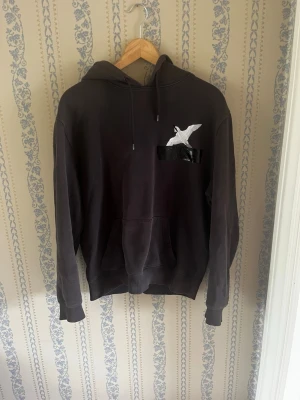 Mörkgrön hoodie från Axel Arigato - Säljer en mörkgrön hoodie med huva och dragsko. På bröstet finns ett vitt fågeltryck ovanför en svart ficka i glansigt material. Hoodien har magficka och ribbade muddar vid ärmar och nederkant. Perfekt för en chill och avslappnad stil.