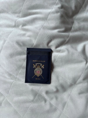 Mörkblå korthållare MORRIS - Snygg mörkblå korthållare i slimmad design med guldfärgat Morris R. Club-emblem och texten M.R.C. på framsidan. Tillverkad i ett glansigt material som ger en exklusiv känsla. Perfekt för kort och ID.