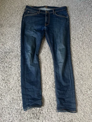 Mörkblå slim fit jeans från Lee - Snygga mörkblå jeans från Lee med raka ben och klassiska kontrastsömmar. Jeansen har fem fickor, knappgylf och Lee-lapp bak i midjan. Tillverkade i slitstark denim med lätt tvättade detaljer för en cool look. Jeansen har två små igensydda hål