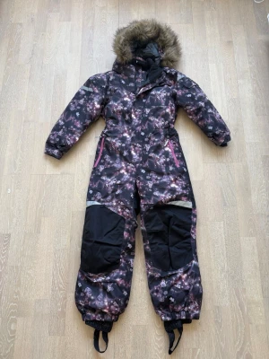 Mörkblommig täckoverall. Stl. 134  - Säljer en mörk täckoverall med blommigt mönster i lila och rosa toner. Overallen har avtagbar huva med fuskpäls, förstärkta knän och reflexdetaljer. Två fickor med dragkedja och snölås vid benslut. Perfekt för kalla vinterdagar.