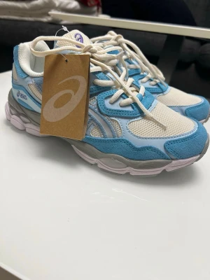 Asics gel-nyc blue - Snygga Asics sneakers i blått och vitt med mesh- och mockadetaljer. Skorna har vita snören, grå sula och klassisk Asics-logga på sidan. Perfekt för dig som gillar sportig och trendig stil. Låg modell med bekväm passform.