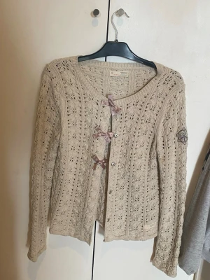 Beige kofta med rosetter Odd Molly - Supersöt beige stickad kofta från Odd Molly med tre ljuslila rosetter framtill. Koftan har ett hålmönster, rund halsringning och långa ärmar. Perfekt för dig som gillar romantisk och feminin stil.anvönd få gånger, står ingen storlek på men hade sagt den passar XS t S 