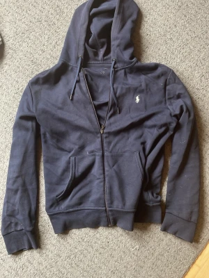 Mörkblå ralph lauren hoodie - Snygg mörkblå hoodie med dragkedja från Polo Ralph Lauren. Klassisk huva med dragsko, två fickor framtill och den ikoniska vita loggan broderad på bröstet. Tillverkad i mjukt bomullsmaterial som är perfekt för en avslappnad stil. Storlekstrycket har försvunnit men det är storlek M!