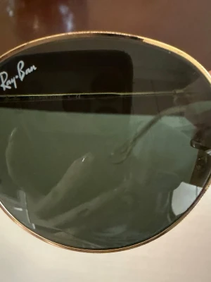 Ray-Ban runda solglasögon guld - Snygga Ray-Ban solglasögon med tunna, runda guldfärgade metallbågar och mörkgröna glas. Klassisk och tidlös modell som ger en cool vibe till din stil. Perfekta för soliga dagar och passar till det mesta.Ray-Ban Round Metal solglasögon med modellnummer RB 3447. Dessa solglasögon har en guldmetallbåge och gröna linser. Storleksinformationen är 50[]21 145 3N. Tillverkade i Italien.
