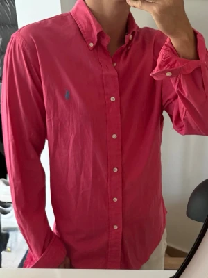 Rosa skjorta med knappar - Säljer en klassisk rosa skjorta med button-down krage och vita knappar. Skjortan har lång ärm och en liten blå logga broderad på bröstet. Perfekt till jeans eller chinos för en clean look.