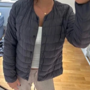 Mörkgrå dunjacka från Uniqlo - Säljer en stilren mörkgrå dunjacka från Uniqlo med rund hals och knappar framtill. Jackan är lätt, quiltad och har en clean look utan krage. Perfekt att slänga på sig när det är kyligt ute. Bilden är lånad men min är bara använd en gång och ser som ny ut. Skriv om fler bilder önskas