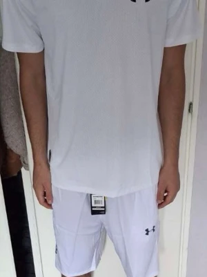Vitt träningsset från Under Armour - Säljer ett fräscht vitt träningsset från Under Armour med t-shirt och shorts. Setet har tryckta loggor och textdetaljer på både tröja och shorts. Perfekt för gym eller löpning. Materialet är lätt och andas bra.