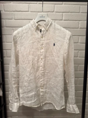 Vit linneskjorta från Polo Ralph Lauren - Klassisk vit skjorta i linne från Polo Ralph Lauren med broderad logga på bröstet. Skjortan har lång ärm, knappar framtill och button-down krage. Perfekt för en stilren och avslappnad look.