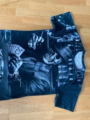Svart t-shirt med dödskalletryck - Cool svart t-shirt med stort tryck av en dödskalle i kostym som spelar poker och röker cigarr. Motiv i gråskala med detaljer som kortlek och flaskor i bakgrunden. Perfekt för dig som gillar unika och edgy plagg.