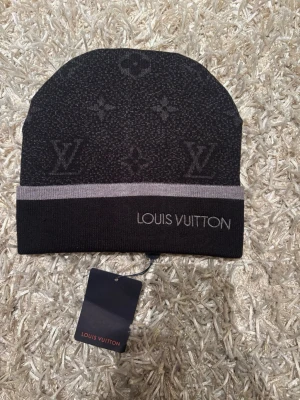 Svart mössa  - Snygg svart mössa med klassiskt monogrammönster och grå rand. Märkets logga är broderad framtill. Perfekt accessoar för att lyfta din stil under kyliga dagar.