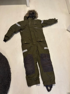 Overall  - Täckoverall i olivgrönt med svarta förstärkningar på knän och bak, reflexdetaljer på armar och ben samt huva med fuskpäls. Praktiska fickor framtill och dragkedja. Perfekt för kalla vinterdagar och utomhusaktiviteter.