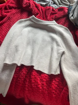Ljusgrå cropped stickad tröja - Säljer en ljusgrå cropped stickad tröja med långa ärmar och ribbad struktur. Tröjan har en hög hals och vågig nederkant som ger en trendig look. Perfekt att styla med höga jeans eller kjol.