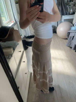Beige långkjol med volangdetaljer - Säljer en beige långkjol med volangdetaljer och bred resår i midjan. Kjolen har ett skirt, lätt material och ett subtilt mönster i ljusare toner. Perfekt för dig som gillar en bohemisk och avslappnad stil.