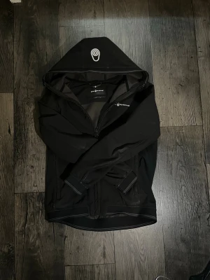 Sail Racing jacka - Snygg svart vindjacka med huva och dragkedja framtill. Jackan har en minimalistisk design med diskret logga på bröstet och justerbara muddar vid ärmsluten. Perfekt för blåsiga dagar och enkel att matcha med olika outfits. 