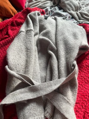 Ljusgrå stickad kofta med knytskärp - Mysig ljusgrå stickad kofta med knytskärp i midjan. Koftan har en avslappnad passform och är perfekt att slänga över en t-shirt eller topp. Materialet känns mjukt och behagligt mot huden. Enkel och stilren design som passar till det mesta.
