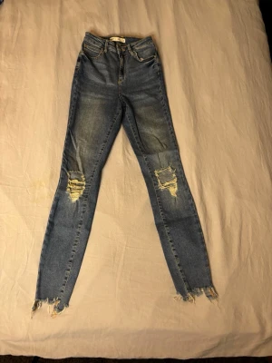 Blå slitna skinny jeans - Säljer ett par blå skinny jeans med hög midja och slitningar på knäna samt fransiga benslut. Jeansen har klassisk femficksdesign och dragkedjegylf. Perfekta för en avslappnad och trendig look.