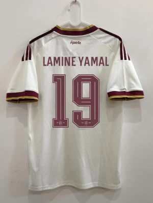 Vit Spanien fotbollströja Lamine Yamal #19 - En vit fotbollströja från Spanien med Lamine Yamal och nummer 19 på ryggen. Tröjan har vinröda detaljer på ärmar och krage samt tre ränder på axlarna. Perfekt för dig som vill stötta landslaget och bära en ikonisk spelare.