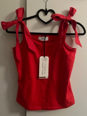 Röd topp med knytband Holly & White - Snygg röd topp köpt från Kappahl med breda axelband som knyts i stora rosetter. I helt nytt skick! Köpt jätte längesen så säljs inte längre.