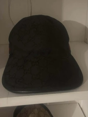 Svart Gucci keps med monogram - Snygg svart keps från Gucci med det klassiska GG-monogrammönstret över hela kepsen. Justerbar rem baktill för perfekt passform. Tillverkad i ett slitstarkt material som känns lyxigt och trendigt.